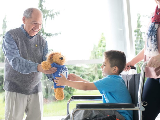 Ein älterer Mann überreicht einem Jungen im Rollstuhl ein Plüschtier des Lions Clubs.